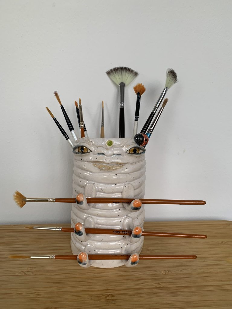 Whimsical gentle white ceramic pot with a caterpillar resemblance. It has little legs holding brushes horizontally and human features such as honey big eyes, wide nose and kind smile