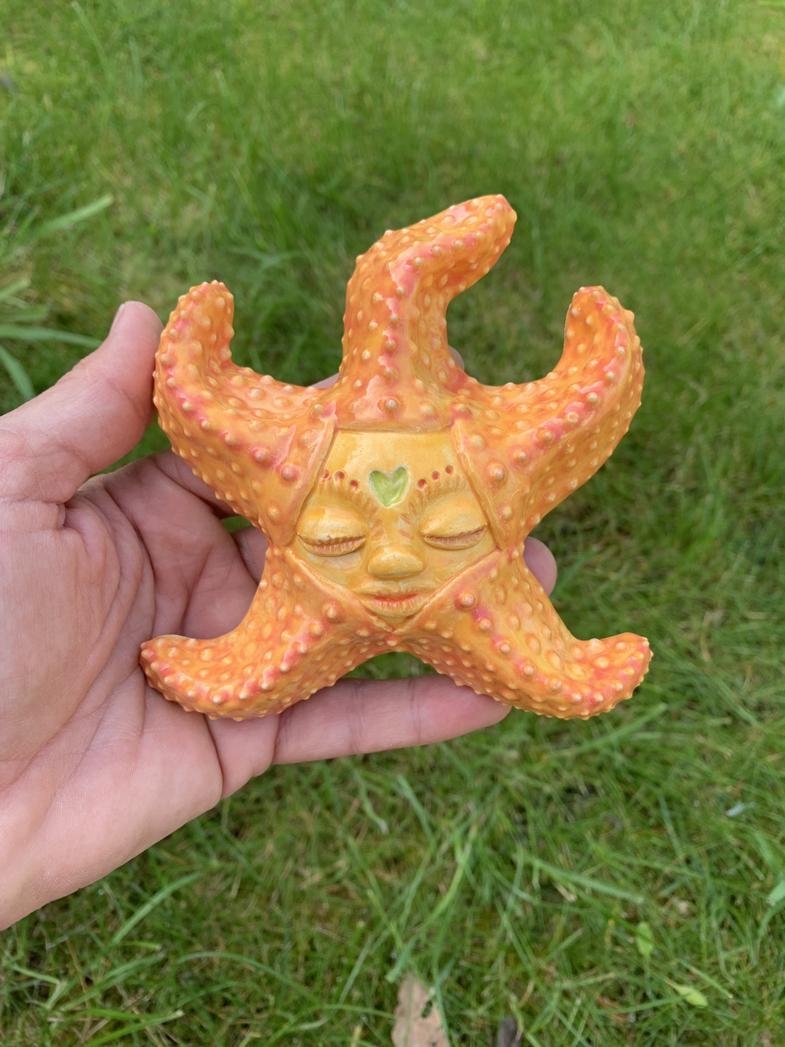 Handmade yellow and orange ceramic starfish sculpture with intricate textured details, closed eyes and a small green heart in the centre of the forehead. The starfish is shown resting in a person's hand outdoors against a soft grassy background, highlighting its size and glossy finish