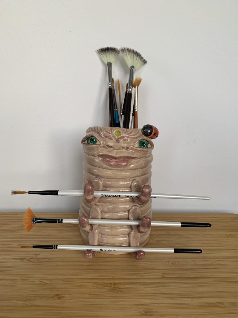 whimsical cream ceramic pot, with resemblance to a caterpillar. Has little legs that hold brushes horizontally. It has female human features like big gentle green eyes, wide nose and big smiley mouth
